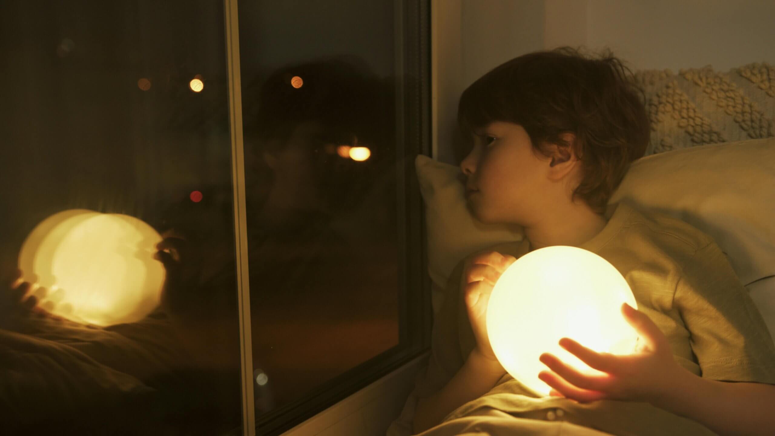 Child laying in bed holding a light-up sensory ball at nighttime.