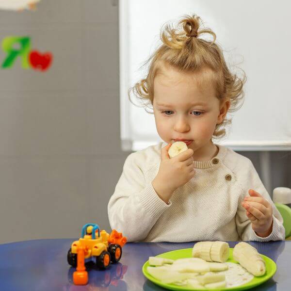Toddler eating a banana in a therapy center with a toy next to them.